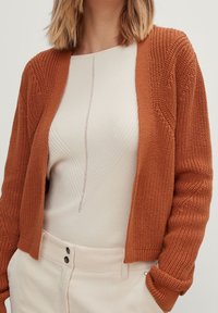 Rustfarbenes Strickjacke mit strukturiertem Strickmuster und offenem Vorderteil, kombiniert mit einem cremefarbenen Oberteil, das subtile vertikale Linien und eine figurbetonte Silhouette aufweist.