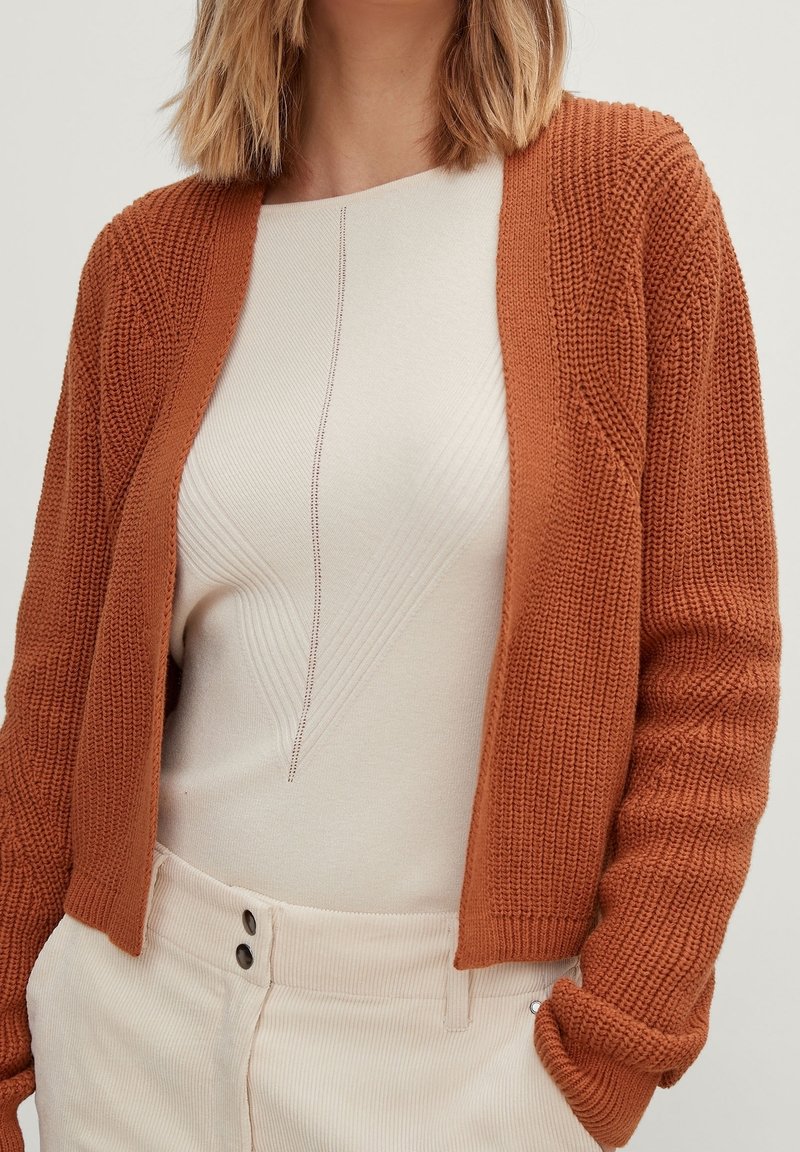 Rustfarbenes Strickjacke mit strukturiertem Strickmuster und offenem Vorderteil, kombiniert mit einem cremefarbenen Oberteil, das subtile vertikale Linien und eine figurbetonte Silhouette aufweist.