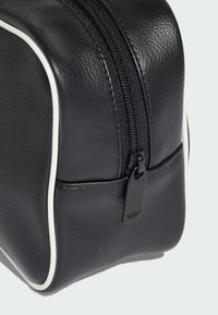 Czarna skórzana kosmetyczka z białymi lamówkami, zamknięciem na suwak i małym suwakiem z logo. Teksturowana powierzchnia i zaokrąglony kształt.