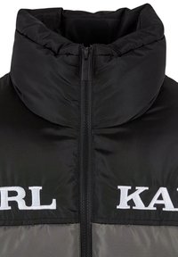 Sort puffergilet med høj krave, der har en central lynlås og hvide broderede logoer "KARL" og "KAT." Glat tekstur, blank overflade.