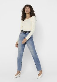 Top de manga larga color crema, vaqueros azules de cintura alta, cinturón negro, tacones blancos, pose de pie contra un fondo blanco liso.