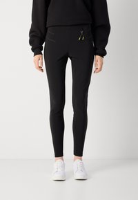 Black fitted leggings with zippered pockets, made from a stretchy material. Paired with white sneakers, featuring a sleek, smooth texture.