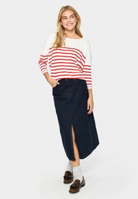 Chemise rayée rouge et blanche à manches longues avec un large décolleté, associée à une jupe bleu marine foncé avec une fente à l'avant et longueur mi-mollet. Mocassins marron portés.