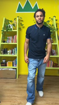 Tamsiai mėlynas polo marškinėlis su mėlynais akcentais, šviesiai mėlynos džinsinės kelnės ir balti sportbačiai. Fone žalia akcentinė siena su lentynomis ir augalais.