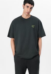 T-shirt noir oversized en coton, avec un col rond et un petit logo vert sur la poitrine. Forme équilibrée avec des manches courtes.