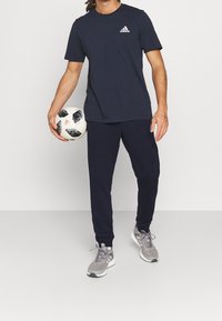 Marinblå sportt-shirt med vitt Adidas-logotyp, parat med matchande marinblå träningsoverallsbyxor. Grå sportskor. Modellen håller en svart och vit fotboll.