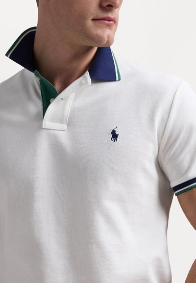 Vit polo skjorta med en marinblå och grön randig krage, med en liten marinblå logotyp på bröstet och en texturerad tyg.