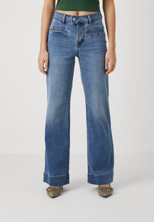 Femme portant un jean bleu taille haute à jambes larges avec fermeture éclair et bouton devant, associé à un top court vert et des chaussures beiges pointues.