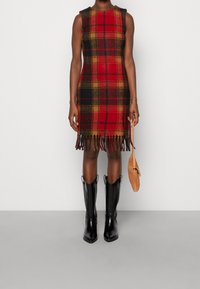 Robe en laine à carreaux rouges et noirs avec un ourlet à franges, sans manches, associée à des bottes de cowboy noires et un sac à main beige.