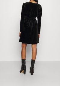 Svart sammetklänning med långa ärmar, bälte i midjan och knälång längd. Bärs med svarta klackboots. Slät textur, minimalistisk design.