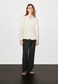 Frau mit langen Haaren, die ein cremefarbenes, geknöpftes Blazer, schwarze weit geschnittene Hose und schwarze Sandalen trägt und vor einem Hintergrund aus weißem und dunklem Boden steht.