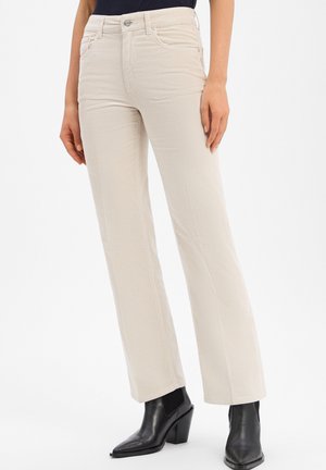 Femme portant un pantalon en velours côtelé beige et des bottines noires, se tenant devant un fond blanc uni.