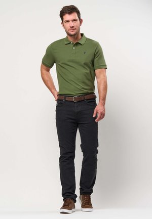 Uomo che indossa una polo verde oliva, jeans neri, cintura marrone e sneakers marroni, in piedi con una mano sui fianchi su uno sfondo semplice.