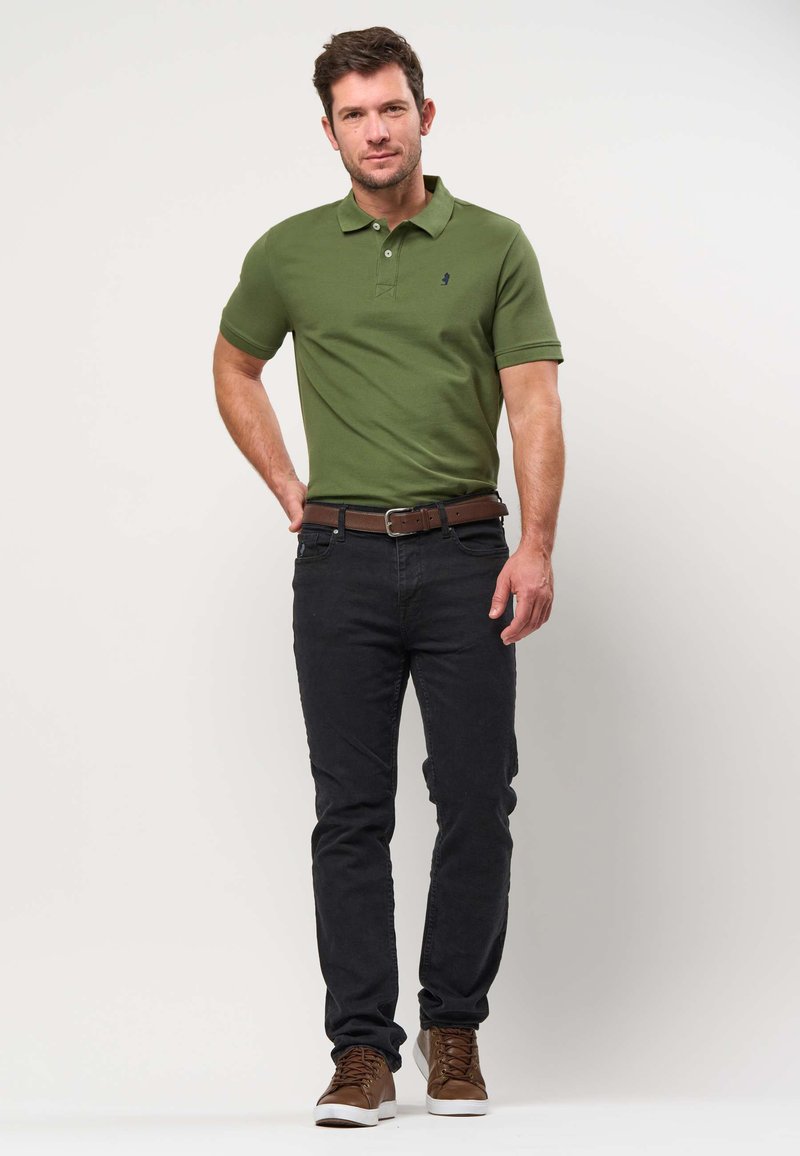 Bărbat purtând tricou polo verde măsliniu, blugi negri, curea maro și adidași maro, stând cu o mână în șold, pe un fundal simplu.