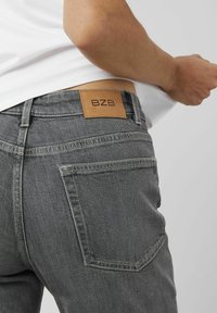 Jean en denim gris avec un patch en cuir BZB à la taille arrière, présentant des détails cousus et un design classique à cinq poches.