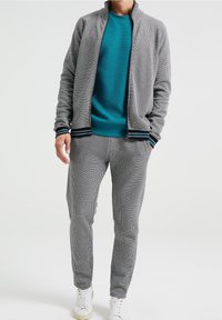 Grå texturerad träningsdräkt med en zip-up jacka och matchande byxor. Jackan har randiga muddar; undertröjan är teal. Vita sneakers.