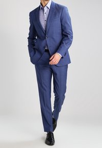 Men's blue suit made of textured fabric, featuring a two-button blazer and flat-front trousers, paired with a light patterned shirt and black shoes.