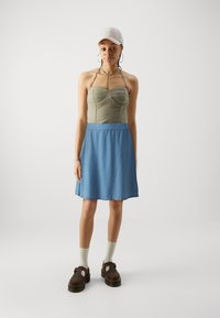 Zöld, bordázott ujjú tanktop, szív alakú dekoltázzsal, kék farmer szoknyával párosítva; barna cipővel és világos zoknival viselve, valamint bézs sapkával.
