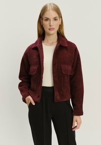 Bordeaux fleecejack met een kraag, twee voorkruiszakken en knoopsluiting, gedragen over een licht geribbeld topje en zwarte broek.
