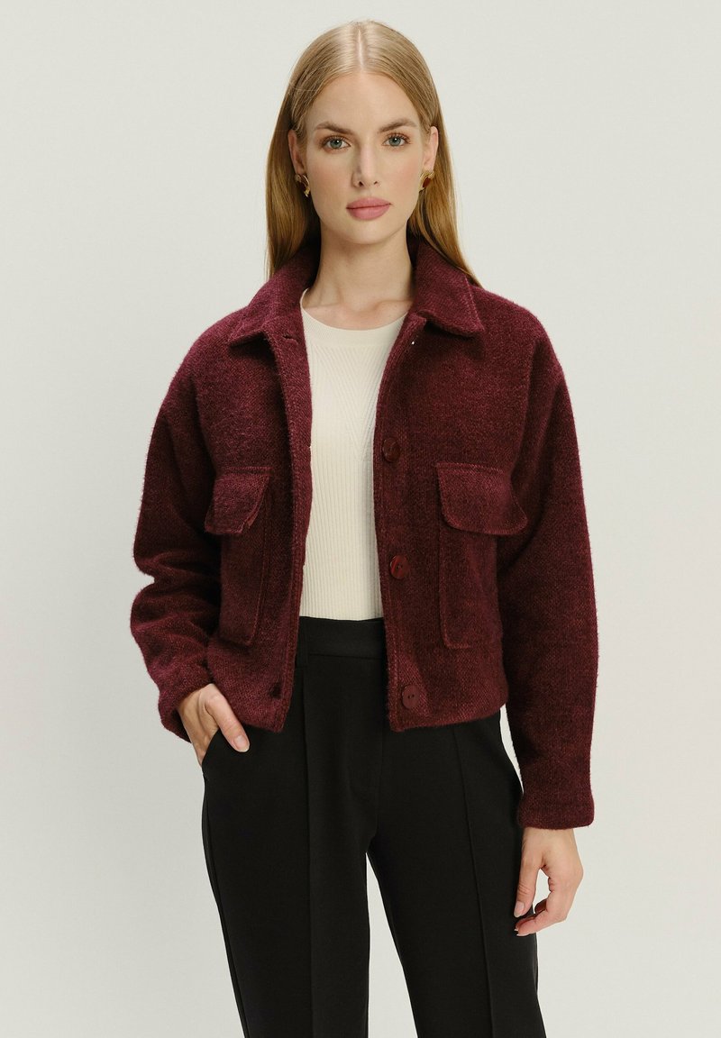 Bordeaux fleecejack met een kraag, twee voorkruiszakken en knoopsluiting, gedragen over een licht geribbeld topje en zwarte broek.