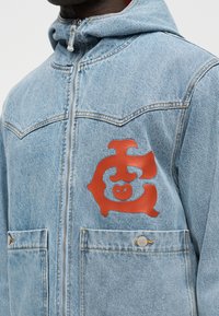 Vyras dėvintis šviesiai mėlyną džinsinę striukę su raudonu abstrakčiu logotipu kairėje krūtinės pusėje, priekyje yra užtrauktukas ir du kišenės su sagomis.