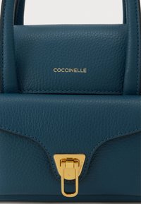 Grønblå læderhåndtaske med struktureret overflade, der har spænde i guld og præget mærkenavn "COCCINELLE" foran.