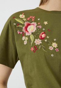 T-shirt verde oliva a maniche corte con ricami floreali in rosa, rosso e giallo. Presenta cuciture intricate e un collo a giro.