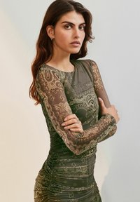 Olívovozelené šaty s priehľadnými dlhými rukávmi, zložitým paisley vzorom, priliehavým strihom a naberaním v oblasti pása. Elegantné a textúrované.