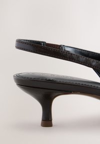 Mørkebrune læder kitten heel sandaler med tynd ankelrem og afrundet lav hæl på en lys baggrund.