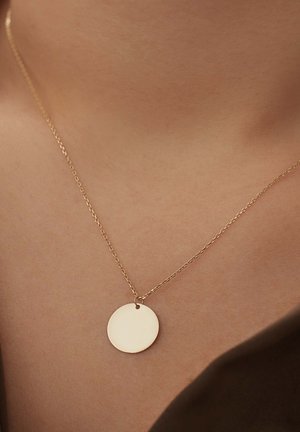 Goldene Halskette mit einem einfachen runden Anhänger, der eine glatte Oberfläche aufweist, hängt an einer zarten Kette vor einem hautfarbenen Hintergrund.