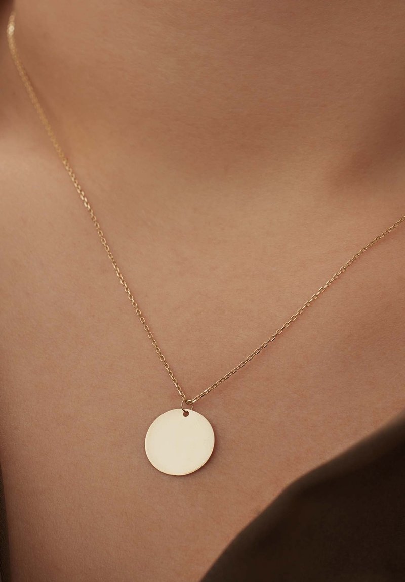 Collier en or avec un pendentif circulaire simple, présentant une surface lisse, suspendu à une chaîne délicate sur un fond de couleur peau.