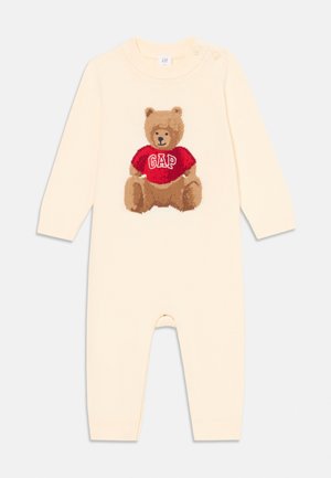 Cremfarbener Baby-Overall mit langen Ärmeln, der auf der Vorderseite einen braunen Teddy-Bär in einem roten GAP-Pullover zeigt und Knöpfe an der Schulter hat.