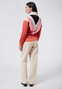 Färgblockerad sweatshirt i korall, rosa och svart med en triangulär design på baksidan. Kombinerad med beige avslappnade byxor och svarta skor.