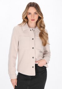 Light beige button-up jacket with two chest pockets and black buttons. Made of soft fabric, featuring a collared neckline and long sleeves.