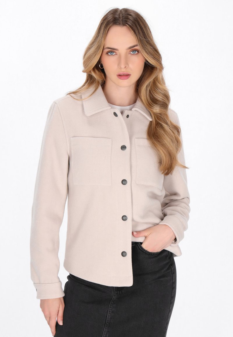 Chaqueta bottona de color beige claro con dos bolsillos en el pecho y botones negros. Fabricada en tejido suave, presenta un cuello con solapa y mangas largas.
