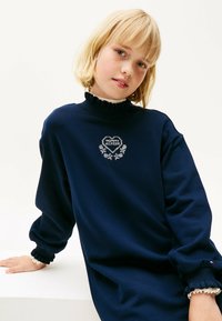 Tmavomodré šaty so svetrom a volánovými límcom a manžetami. Obsahujú grafiku v tvare srdca s nápisom "MOMMY HILFIGER" a kvetinovými akcentmi.