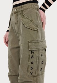 Pantaloni de tip cargo în nuanță de verde măsliniu, confecționați dintr-un material durabil, având multiple buzunare și inscripții negre pe lateral. Un top cu dungi iese în evidență deasupra.