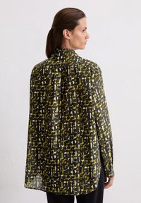 Femme aux cheveux bruns portant un haut à manches longues à motifs noir, vert olive et blanc, se tenant de profile contre un mur blanc uni.