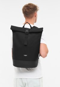 Sac à dos noir à rabat fabriqué en tissu doux avec une finition élégante, doté d'une sangle frontale et d'une étiquette de marque près de la base.
