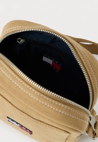 Béžová ľadvinka z plátennej látky so zipsovým uzáverom, vnútornej vreckom, bielym prešívaním a značkovou štítkom s modrými, bielymi a červenými pruhmi.