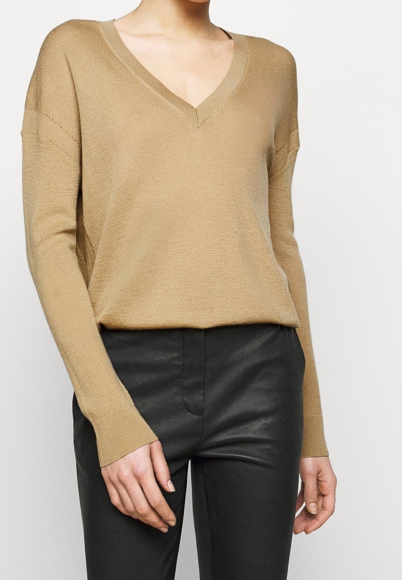 Femme portant un pull beige à col en V et manches longues, rentré dans un pantalon en cuir noir taille haute, visible du cou jusqu’aux hanches.