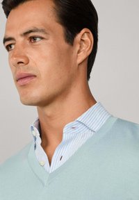 Svetlo modrý sveter s výstrihom do V cez bielu a modrú pruhovanú košeľu s golierom. Hladká textúra, obsahuje gombíkový golier a jemné detaily šitia.