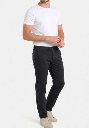 Hombre con camiseta blanca lisa de manga corta, pantalones negros y zapatillas beige claras, de pie frente a un fondo blanco.