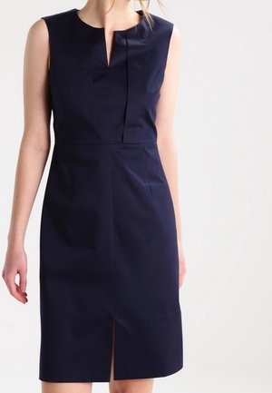 Mouwloze, marineblauwe jurk tot op de knie met een kleine voorste split en een bescheiden V-vormige inkeping in de hals, gedragen door een staand persoon.