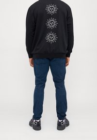 Sudadera negra con tres gráficos de sol blancos en la parte posterior, combinada con jeans azules y zapatillas deportivas negras. Tela suave, corte relajado.