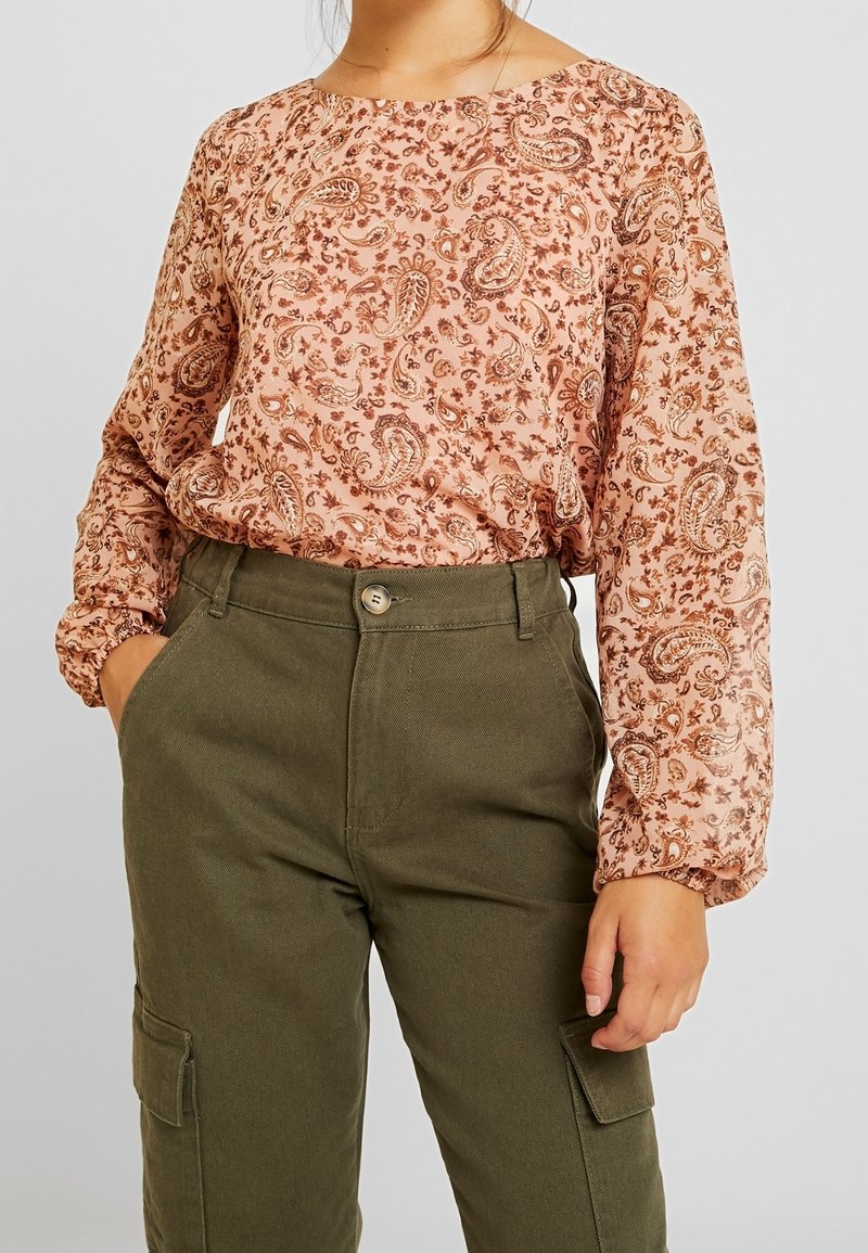 Personne portant un chemisier rose à manches longues avec motif cachemire rentré dans un pantalon cargo taille haute vert olive avec des poches avant.