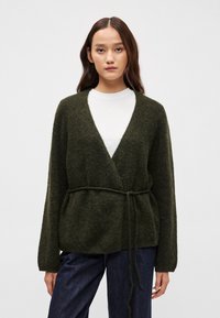 Tmavě zelený kardigan s obálkovým střihem, strukturovaným povrchem, dlouhými rukávy a uvázaným páskem. Nosený přes bílý top a kombinovaný s tmavými džíny.