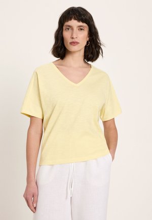 Woman with shoulder-length dark hair wearing gold hoop earrings, a pale yellow V-neck T-shirt, and white pants, standing with one hand in pocket.