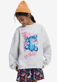 Grå sweatshirt med oversized passform, prydd med en färgglad Stitch-grafik. Matchas med en mönstrad mörk kjol med frukt- och blommönster. Orange mössa.