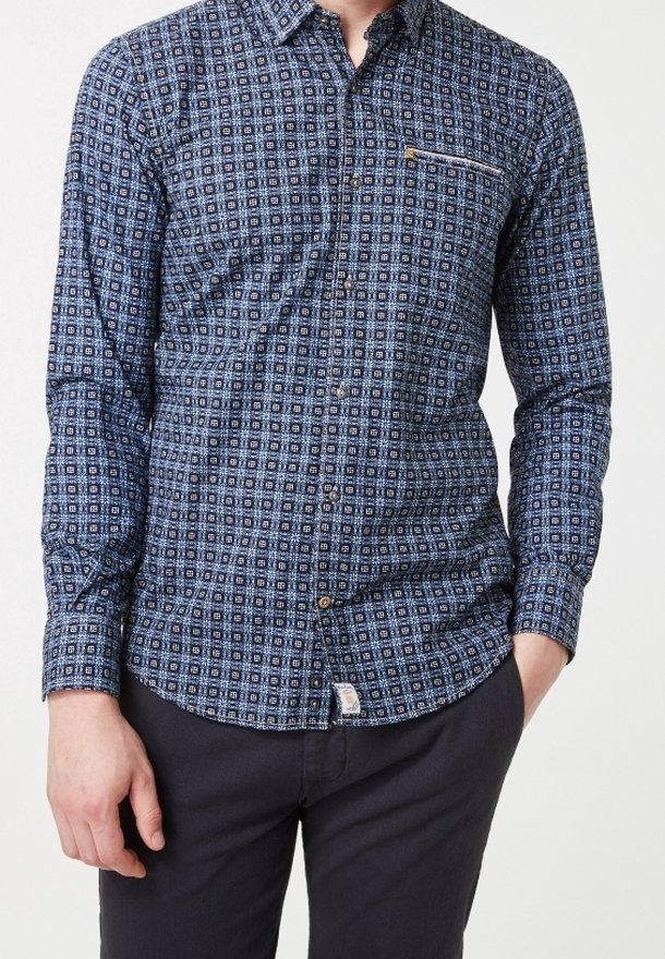 Homme portant une chemise à boutons à manches longues bleue à motifs avec une poche zippée sur la poitrine et un pantalon foncé, debout avec une main dans la poche.